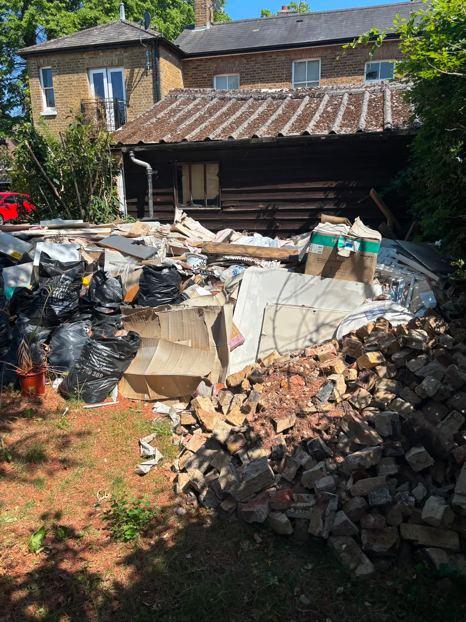 Rubble, plasterboard, cardboard and bagged builders waste piled against a timber outbuilding.