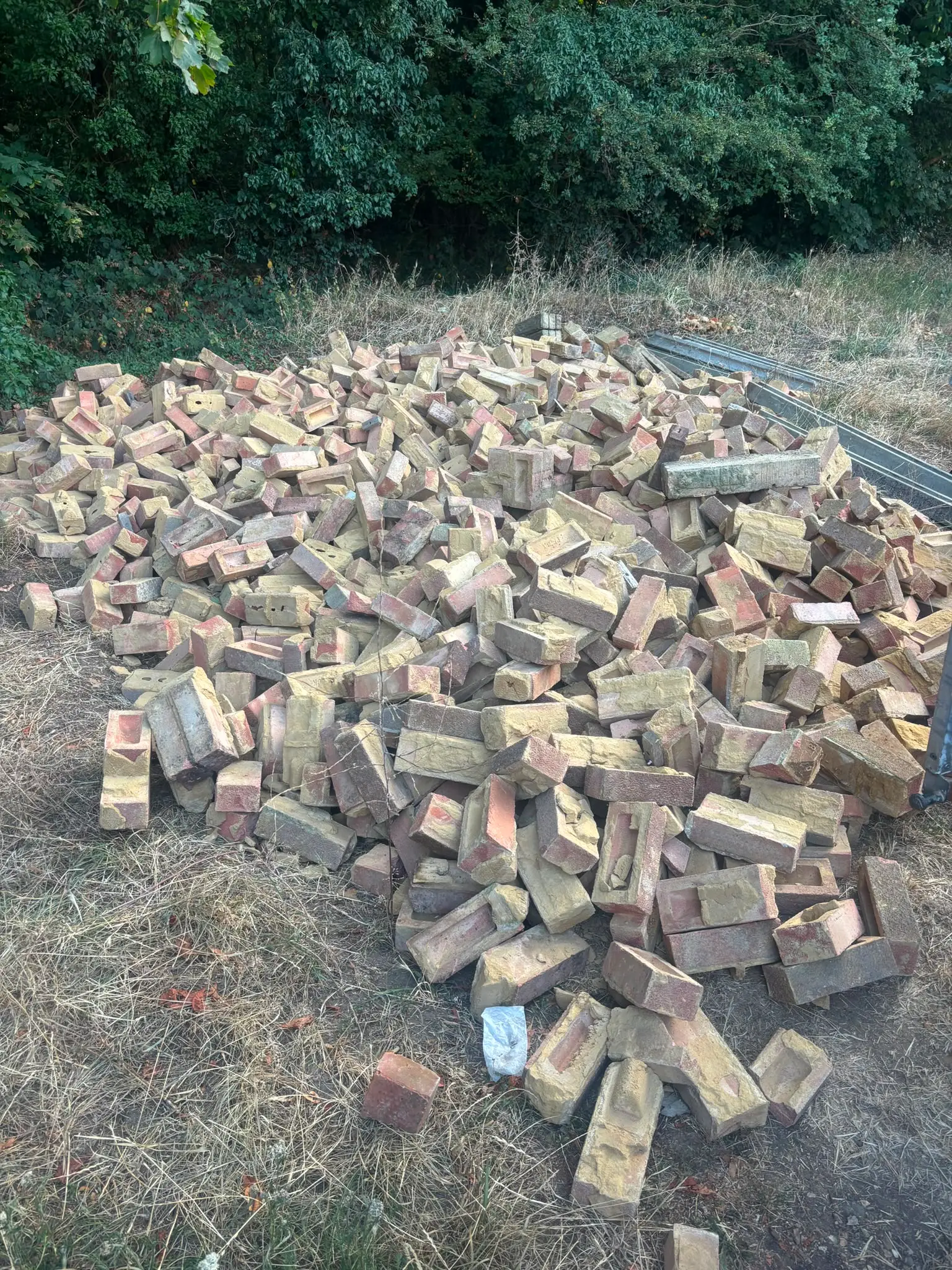 Large pile of reclaimed bricks and metal beams left on grass beside a hedge before removal.